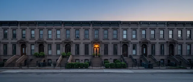 A row of matching brownstone townhouses at blue hour, one lamp lit over one front door