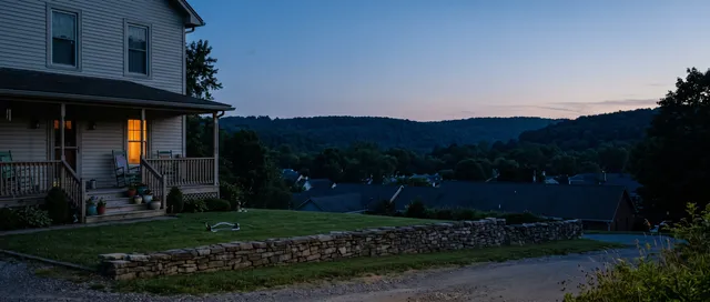 A porch on a hillside street above a small town at blue hour, one front window lit